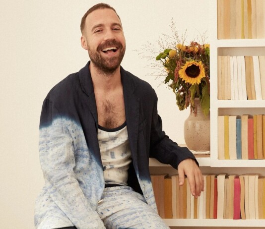 Queer Designer Louis-Gabriel Nouchi Brings High Fashion to OnlyFans Man with a beard laughs while sitting beside a bookshelf with a bouquet of sunflowers in a vase behind him.