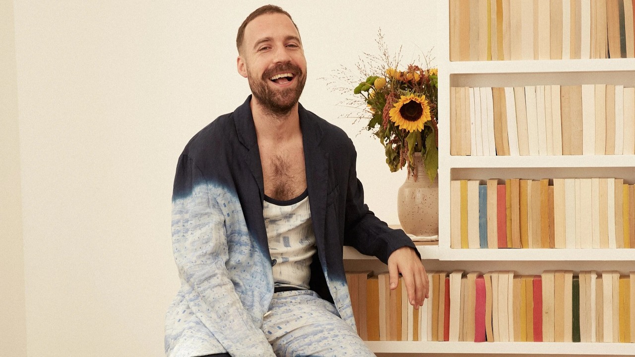 Man with a beard laughs while sitting beside a bookshelf with a bouquet of sunflowers in a vase behind him.