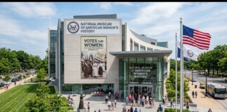 Front view of the National Museum of American Women’s History with large banner and American flag outside; people gather at the entrance on a sunny day.