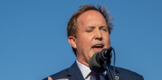 Man in a blue suit delivering a speech at a microphone outdoors against a clear blue sky?