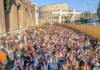 Huge crowd of people gathered outdoors at a festival or concert near the Colosseum in Rome, with yellow barriers along the right side and clear blue sky above.