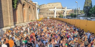 Huge crowd of people gathered outdoors at a festival or concert near the Colosseum in Rome, with yellow barriers along the right side and clear blue sky above.