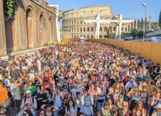 Huge crowd of people gathered outdoors at a festival or concert near the Colosseum in Rome, with yellow barriers along the right side and clear blue sky above.