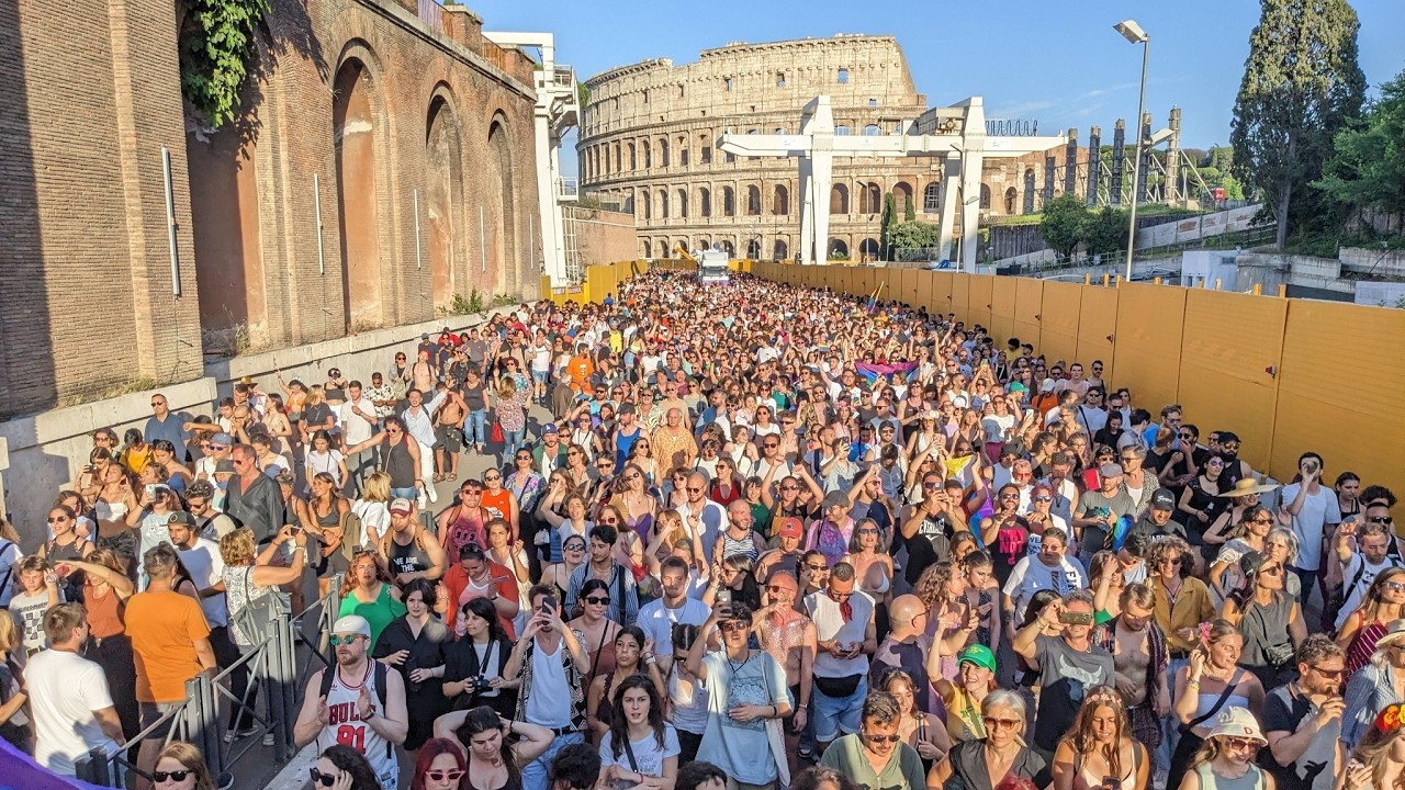 Huge crowd of people gathered outdoors at a festival or concert near the Colosseum in Rome, with yellow barriers along the right side and clear blue sky above.