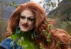 Drag queen with bright red wig and bold makeup posing outdoors among trees, wearing a green jacket and turquoise earrings.