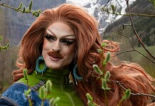Patagonia Sues Drag Climate Activist Pattie Gonia in Trademark Dispute Drag queen with bright red wig and bold makeup posing outdoors among trees, wearing a green jacket and turquoise earrings.