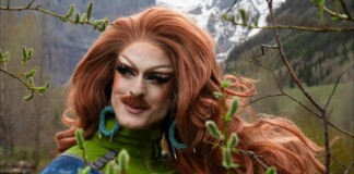 Drag queen with bright red wig and bold makeup posing outdoors among trees, wearing a green jacket and turquoise earrings.