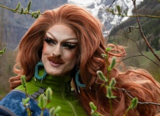 Drag queen with bright red wig and bold makeup posing outdoors among trees, wearing a green jacket and turquoise earrings.
