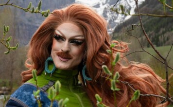 Drag queen with bright red wig and bold makeup posing outdoors among trees, wearing a green jacket and turquoise earrings.