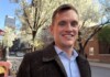 Smiling man in a brown tweed coat on a springtime city street with blossoming trees.