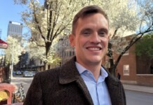 Smiling man in a brown tweed coat on a springtime city street with blossoming trees.
