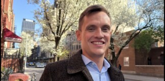 Smiling man in a brown tweed coat on a springtime city street with blossoming trees.