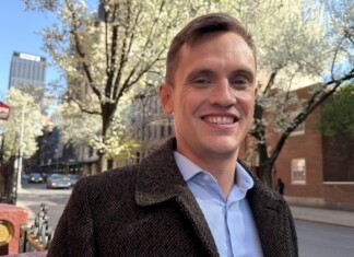 Out Gay Carl Wilson Wins NYC Stonewall District Council Race, Beating Mamdani-Endorsed Candidate Smiling man in a brown tweed coat on a springtime city street with blossoming trees.