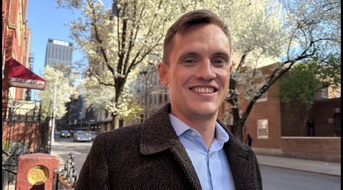 Smiling man in a brown tweed coat on a springtime city street with blossoming trees.