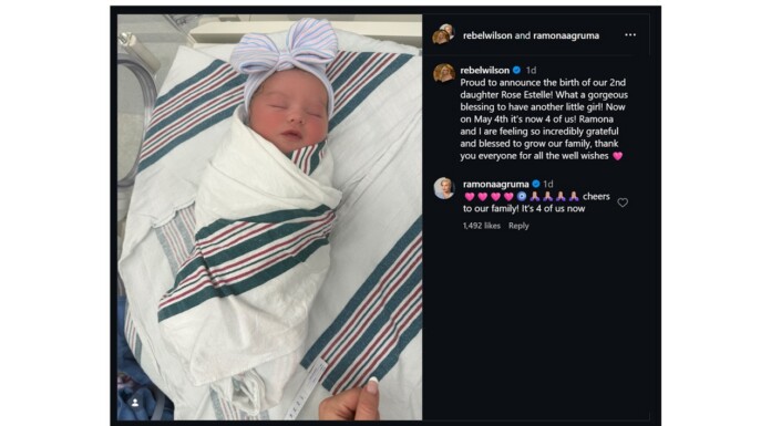 Rebel Wilson and Wife Ramona Agruma Welcome Second Daughter, Rose Estelle Newborn baby swaddled in a striped hospital blanket with a pink bow headband, lying in a hospital bed. That the image shows a birth announcement in a social post context.