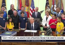 Man in a dark suit sits at a large wooden desk in a formal room, surrounded by adults and children wearing blue, yellow, and red shirts with flags in the background.