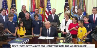 Man in a dark suit sits at a large wooden desk in a formal room, surrounded by adults and children wearing blue, yellow, and red shirts with flags in the background.