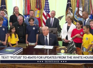 Man in a dark suit sits at a large wooden desk in a formal room, surrounded by adults and children wearing blue, yellow, and red shirts with flags in the background.