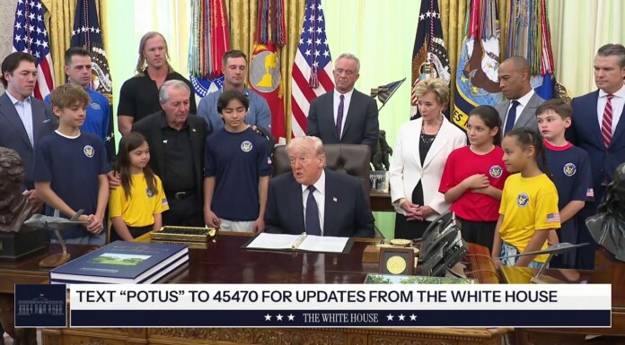 Trump Targets Trans Athletes While Speaking to Children in Oval Office Man in a dark suit sits at a large wooden desk in a formal room, surrounded by adults and children wearing blue, yellow, and red shirts with flags in the background.