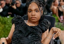 Portrait of a woman in a black, ruffled evening gown seated in a wheelchair at a red carpet event, with photographers in the background.