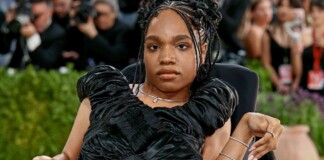 Portrait of a woman in a black, ruffled evening gown seated in a wheelchair at a red carpet event, with photographers in the background.