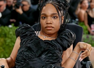 Portrait of a woman in a black, ruffled evening gown seated in a wheelchair at a red carpet event, with photographers in the background.