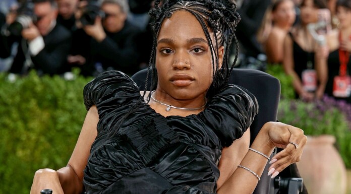 Portrait of a woman in a black, ruffled evening gown seated in a wheelchair at a red carpet event, with photographers in the background.