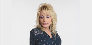 Portrait of a blonde woman with pink and purple makeup wearing a dark patterned blouse, looking downward toward her left shoulder.