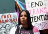 A speaker at a protest talks into a microphone, with signs reading 'Free Them All' and 'End Trans Detentions' behind her.