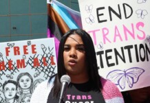 A speaker at a protest talks into a microphone, with signs reading 'Free Them All' and 'End Trans Detentions' behind her.