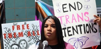 A speaker at a protest talks into a microphone, with signs reading 'Free Them All' and 'End Trans Detentions' behind her.
