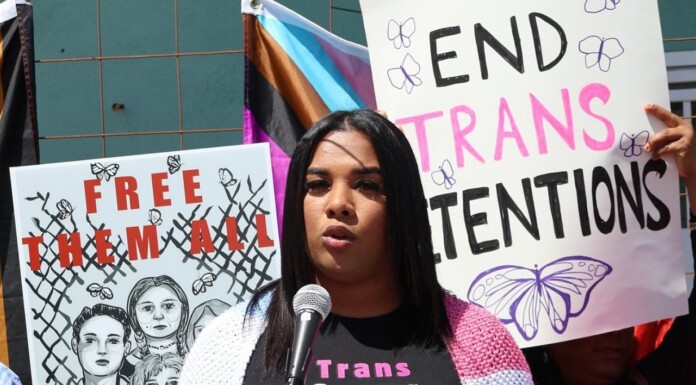 A speaker at a protest talks into a microphone, with signs reading 'Free Them All' and 'End Trans Detentions' behind her.
