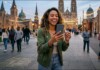 Woman smiling while recording with her smartphone in a lively city square with string lights and church spires behind her at dusk.