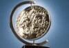 Shiny silver coin mounted in a curved metal stand against a blue sky background, seen from the side.