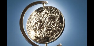 Shiny silver coin mounted in a curved metal stand against a blue sky background, seen from the side.