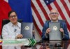 Two officials sit at a desk, holding signed folders with government seals, in front of a flag backdrop (US flag and others).