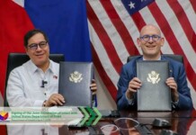 Two officials sit at a desk, holding signed folders with government seals, in front of a flag backdrop (US flag and others).