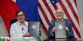 Two officials sit at a desk, holding signed folders with government seals, in front of a flag backdrop (US flag and others).