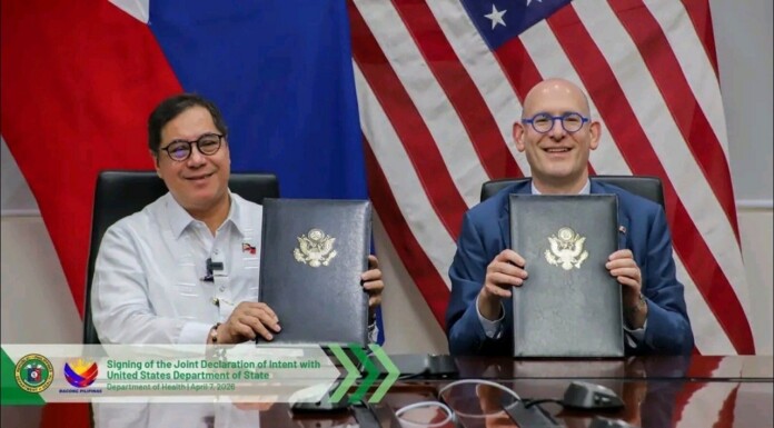 Two officials sit at a desk, holding signed folders with government seals, in front of a flag backdrop (US flag and others).