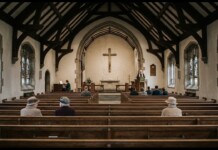 Inside a church with wooden pews and dark beams, a preacher stands at the pulpit near the illuminated altar cross.