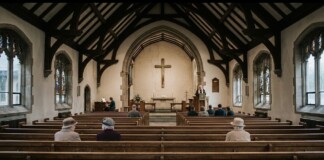 Inside a church with wooden pews and dark beams, a preacher stands at the pulpit near the illuminated altar cross.