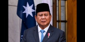 Formal portrait of a man in a dark suit and light blue tie, wearing a medal on his lapel, indoors with a flag in the background.