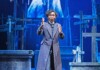 Actor in a long striped coat on a stage with wooden crosses, gesturing dramatically.