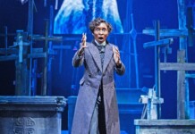 Actor in a long striped coat on a stage with wooden crosses, gesturing dramatically.