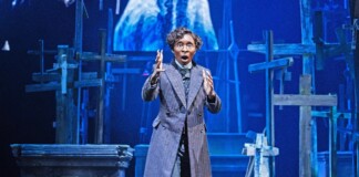 Actor in a long striped coat on a stage with wooden crosses, gesturing dramatically.