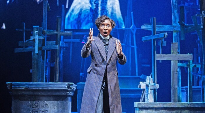 Actor in a long striped coat on a stage with wooden crosses, gesturing dramatically.
