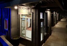 Nighttime exterior of a storefront labeled Nordheim Court along a tiled sidewalk with a blue-lit wall to the left.