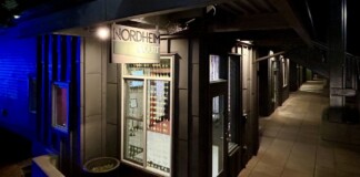 Nighttime exterior of a storefront labeled Nordheim Court along a tiled sidewalk with a blue-lit wall to the left.