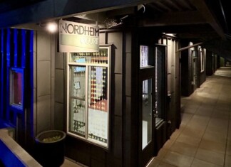 Nighttime exterior of a storefront labeled Nordheim Court along a tiled sidewalk with a blue-lit wall to the left.