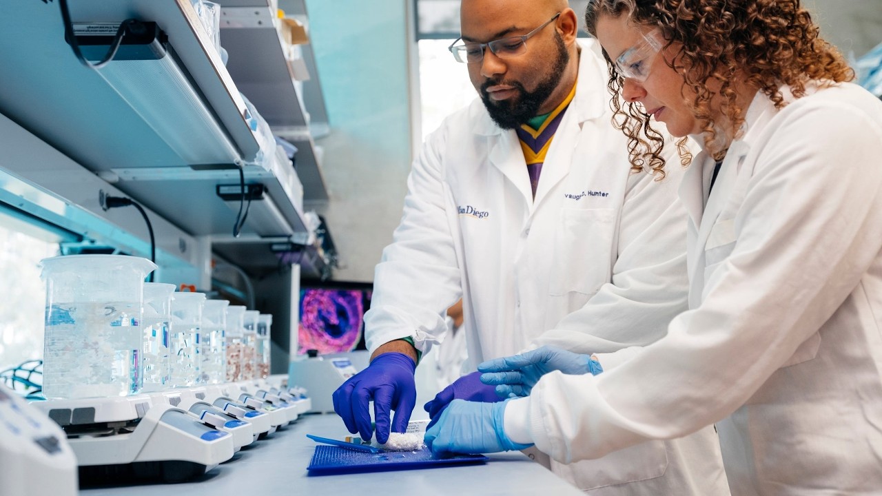 Scientists in a lab coat duo handling a sample on a blue mat, surrounded by analytical equipment and beakers in a modern lab.